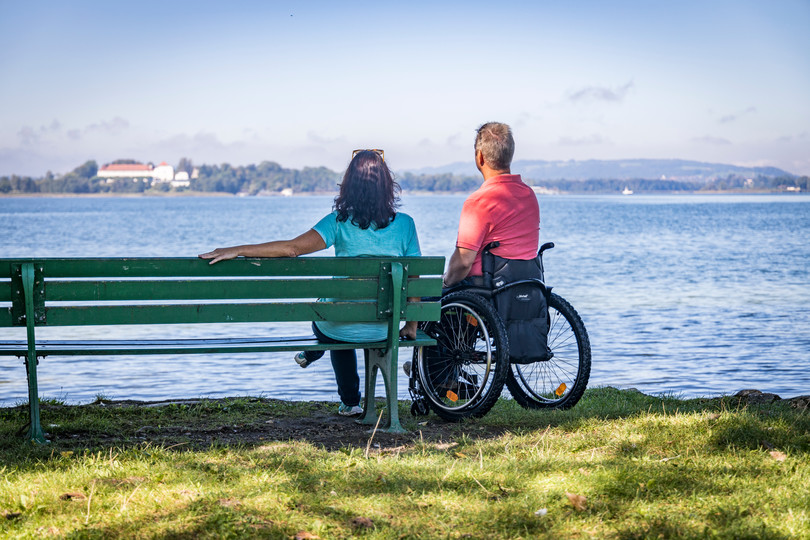 Eine Frau sitzt auf einer Bank und ein Mann im Rollstuhl steht neben ihr, beide blicken auf den Chiemsee.