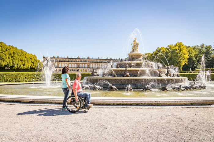 Ein Rollstuhlfahrer und eine Frau vor dem großen Latonabrunnen des Schloss Herrenchiemsees bei Sonnenschein.
