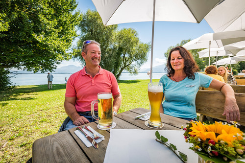 Ein Mann im Rollstuhl und eine Frau im Biergarten bei Sonnenschein mit kühlem Bier auf dem Tisch.