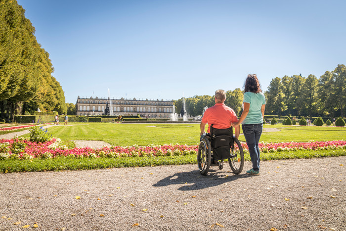 Ein Rollstuhlfahrer und eine Frau vor der Parkanlage des Schloss Herrenchiemsees. Im Vordergrund sind Blumen gepflanzt, im Hintergrund ist das Schloss zu sehen.