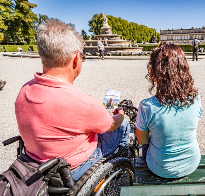 Rollstuhlfahrer und eine Frau sitzen auf einer Bank mit Blick auf das Schloss und Barrierefrei Broschüre in der Hand